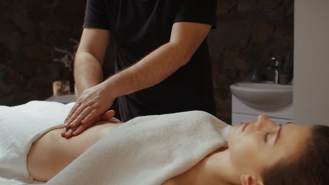 Male masseur hands moves slow burning fat by anti-cellulite massage. Young woman receiving stomach massage at spa salon, lying on massage table slide dolly camera movement