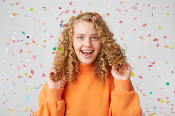 A magical time. Portrait of a very happy girl in orange sweater touches plays with her curly hair, smiling at confetti falling