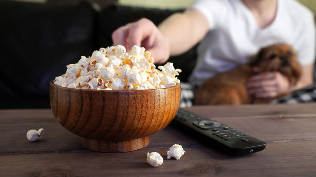 Wooden bowl with salted popcorn and TV remote on wooden table. In the background, a man with a red dog watching TV on the couch