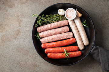 Assorted fresh raw sausages with thyme, rosemary and garlic in a cast iron pan on a dark concrete table. Top view with copy space. Flat lay