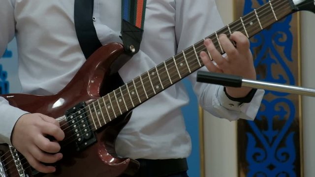 Guy pushes strings with a pick and fingers clamps frets on a solo guitar. A teenager in a white shirt plays a brown electric guitar. Concert performance. Concept on the topic of musical youth.