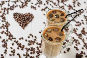 Two glasses of cold coffee on the background of scattered grains in the shape of a heart on a white wooden table.