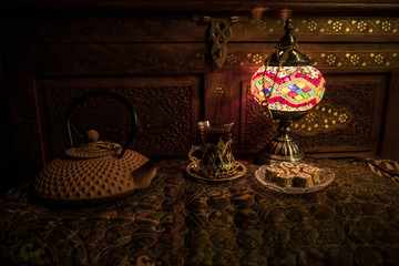 Arabian tea in glass with eastern snacks on vintage wooden surface. Eastern tea concept. Low light lounge interior with carpet. Empty space.