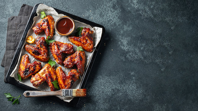 Roasted chicken wings in barbecue sauce with sesame seeds and parsley in a baking tray on a dark table. Top view with copy space. Tasty snack for beer on a dark background. Flat lay