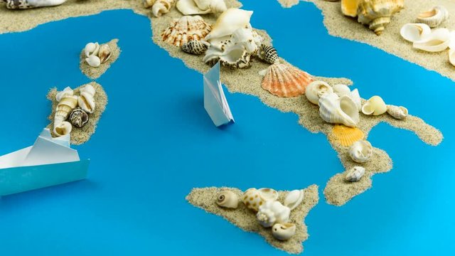 Paper boats coming to the shores of Italy. The map of Italy and the islands is made of sand and seashells.
