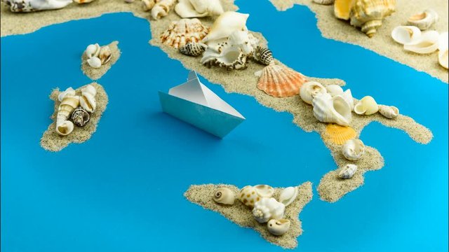 Paper boats coming to the shores of Italy. The map of Italy and the islands is made of sand and seashells.
