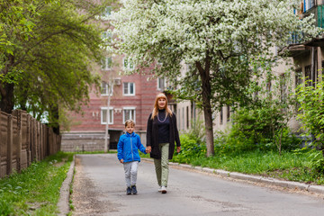 Fototapeta na wymiar a woman of fifty walks by the hand with her seven-year-old grandson in the city quarter in the spring, blooming Apple trees