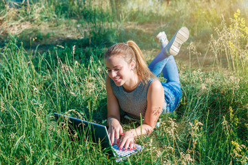 A young girl communicates with her beloved through a computer in the open air lying on the green grass.