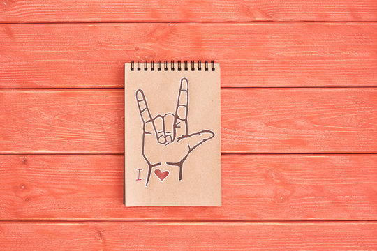 Notebook of Kraft paper with a painted symbol ASL (American Sign Language) ILY (I Love you) that lies on the textured wooden plank of the color Living Coral, flat lay, top view, copy space