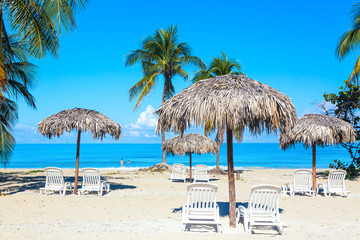 A sun loungers under umbrellas on the sandy beach with palms by the sea and sky. Vacation background. Idyllic beach landscape.
