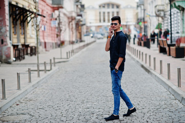 Stylish indian model man in casual clothes, black shirt and sunglasses posed outdoor at street of India and speaking at telephone.
