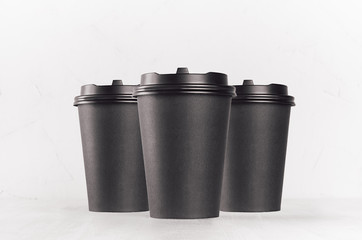 Coffee mockup - group of three blank black paper cups with cap closeup on white wood table, coffee shop interior. Modern elegant concept for branding identity, advertising, design.