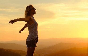 Fototapeta na wymiar Happy woman with open arms stay on the peak of the mountain cliff edge under sunset light sky enjoying the success, freedom and bright future