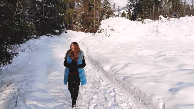 Young beautiful girl walking in the snowy winter forest. She is happy and calm. The girl looks on the sides. The sun is shining. Shooting from the quadcopter. Slow motion.