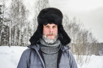 Siberian Russian man with a beard in hoarfrost in freezing cold in the winter freezes in a village in a snowdrift and wears a hat with a earflap.