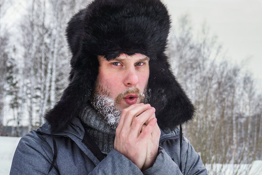 a frozen man with a beard with weathered skin and lips in the winter in a forest with snowdrifts warms his hands with the cold from a walk after walking through the village