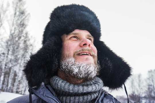 Happy Siberian Russian man with a beard smiles and lets out steam from his mouth in hoarfrost in freezing cold in the winter freezes in a village in a snowdrift and wears a hat with a earflap.