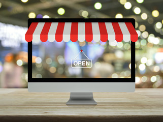 Desktop modern computer monitor with online shopping store graphic and open sign on wooden table over blur light and shadow of shopping mall, Business internet shop online concept