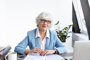 Attractive gray haired senior businesswoman in fashionable suit and glasses working in her office,...