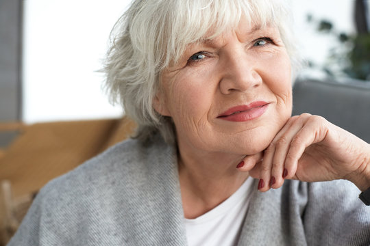 Close up image of adorable charming female pensioner with white short hair, wrinkles and wise blue eyes relaxing at home, having happy look, looking at camera. Beauty, style and maturity concept