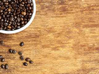 Macro shot of coffee beans in ceramic bowl and scattered coffee seeds on wooden background. Flat lay, copyspace