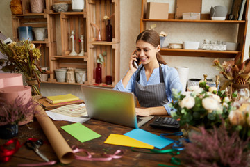 Portrait of female small business owner using laptop and speaking by phone in flower shop, copy space