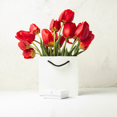 White Gift Bag, small white gift box, petals and bouquet of red tulips on a light stone background. The concept of an offer for an engagement or a marriage