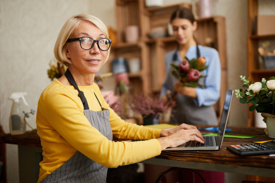 Side View Portrait Of Mature Female Small Business Owner Using Laptop And Looking At Camera In Flower Shop, Copy Space