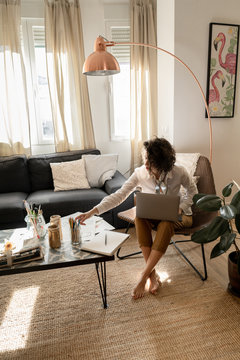 Woman with laptop on chair near low table with pencils, brushes and paper in room