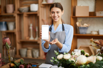 Waist up portrait of smiling female florist holding smartphone with blank screen presenting mobile...