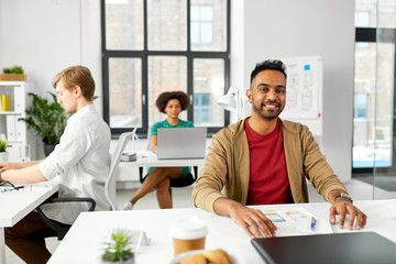 business and people concept - happy smiling indian male creative worker with laptop sitting at office table