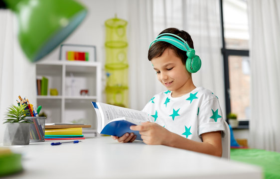 education, childhood and technology concept - happy schoolboy in headphones reading book at home