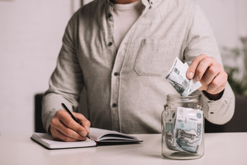 cropped shot of man writing in notebook and putting russian rubles banknotes in glass jar