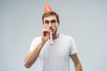 Studio shot of unshaven young Caucasian male blowing whistle while celebrating birthday, having...