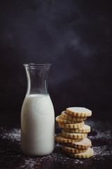 A bottle of milk and a stack of cookies sprinkled with icing sugar, against a dark background