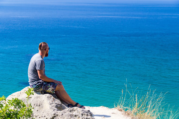 Naklejka na ściany i meble A man in a gray T-shirt sits by the turquoise sea. Beautiful view of the sea.