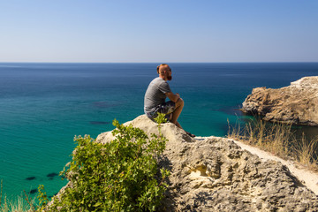 A man in a gray T-shirt sits by the turquoise sea. Beautiful view of the sea.