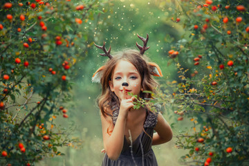 little girl fawn in green summer forest, child plays hide-and-seek holds finger to lips, keeps...