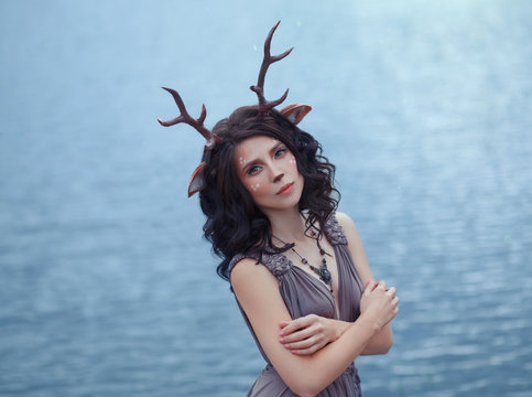 interesting photo of mother deer in front of lagle lake, characters of the fairy forest, image of a sad woman-faun near a large lake, unusual makeup, with homemade horns and ears on dark curly hair