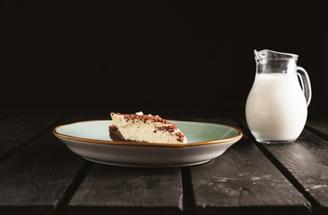 Photo of chocolate bio cheesecake in a retro plate with milk on wooden background. Dark food Photography of sweet and illuminated dessert.