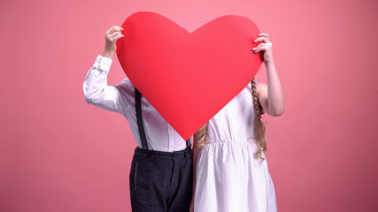 Young boy and girl hiding behind red paper hearts and kissing, Valentines day