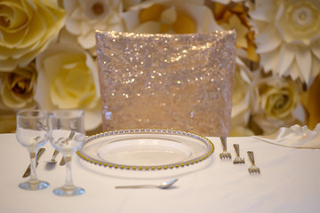 Fototapeta na wymiar Sitting arrangement at a formal event or fine dining restaurant featuring transparent plates with golden details, glassware and silverware in the order of use set against a paper flowers wall