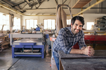 Woodworker smiling while leaning on a bench in his workshop - obrazy, fototapety, plakaty