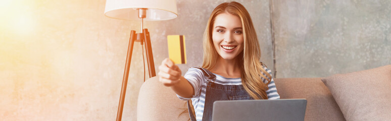 happy beautiful woman showing credit card and sitting on sofa with laptop