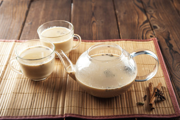 Masala tea on a textural wooden background. A teapot is a transparent tea pot with mugs and Indian national tea masala. Milk, ginger, cardamom, cloves, nutmeg, star anise, cinnamon