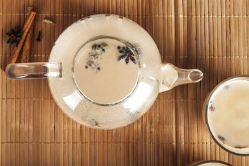 Masala tea on a textural wooden background. A teapot is a transparent tea pot with mugs and Indian national tea masala. Milk, ginger, cardamom, cloves, nutmeg, star anise, cinnamon