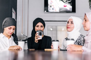 Four girlfriends in hijabs sit at a table in a cafe, drink coffee and communicate with each other. Female friendship
