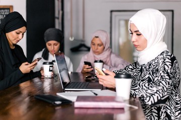 Four girlfriends in hijabs sit at a table in a cafe communicating on social networks through phone. Problems of modern youth