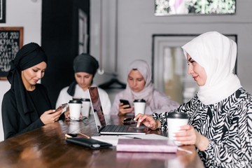 Four girlfriends in hijabs are sitting in a cafe chatting on social networks through the phone. Problems of modern youth