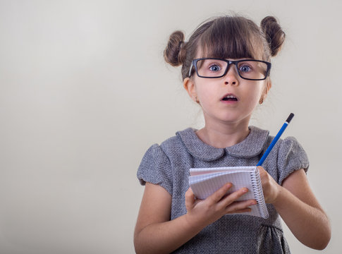 Surprised Cute Child In Eyeglasses,  Writing In Notebook Using Pencil, Keeping Mouth Wide Open. Four Years Old Kid, Isolated On Grey, Space For Advertising Text